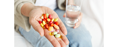 Een vrouw die zit met haar hand uitgestoken. Op haar hand liggen voedingssupplementen in diverse kleuren en vormen. In haar andere hand heeft ze een glas water vast.