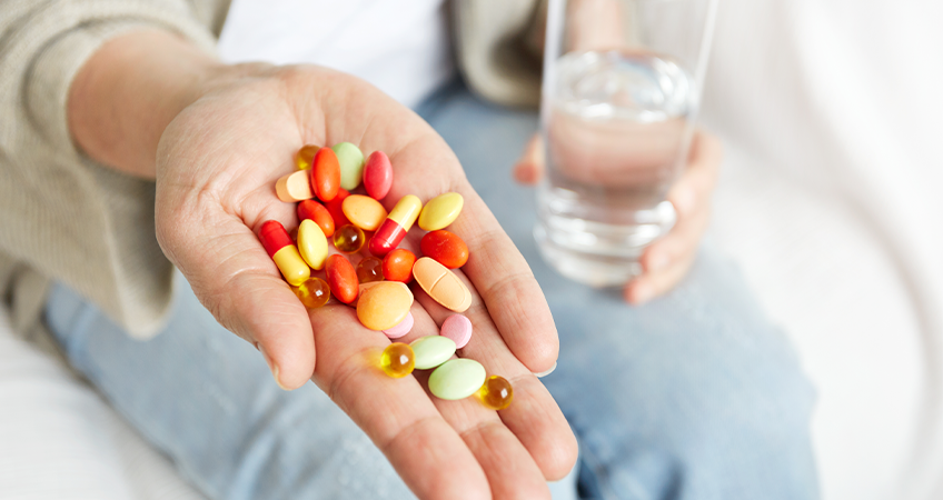 Een vrouw die zit met haar hand uitgestoken. Op haar hand liggen voedingssupplementen in diverse kleuren en vormen. In haar andere hand heeft ze een glas water vast.